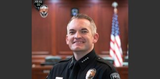 Police officer in uniform smiling in a courtroom with an American flag in the background and badge visible on chest.