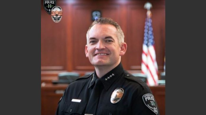 Police officer in uniform smiling in a courtroom with an American flag in the background and badge visible on chest.