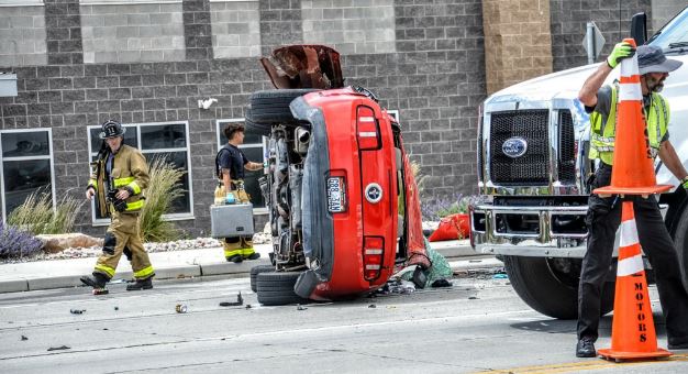 Red car flipped onto its side in the middle of the street, with firefighters, a tow truck, and traffic cones securing the scene amid scattered debris.