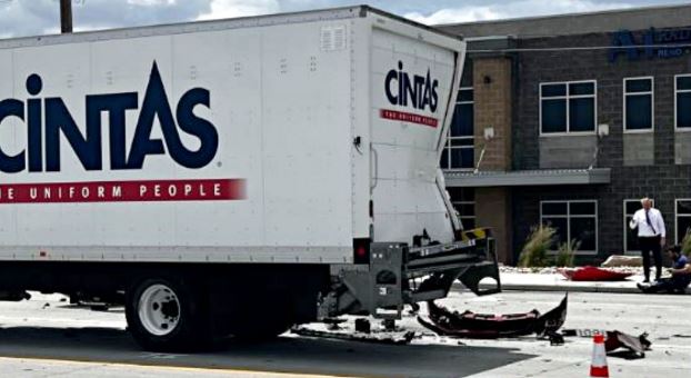 White Cintas delivery truck with rear damage and scattered debris on the street; an orange cone and a man in dress clothes stand near a building in the background.