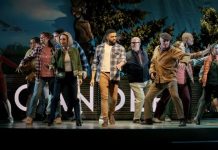 Diverse group of people dancing on a stage, performing a coordinated routine with a colorful backdrop behind them.