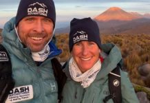 Two hikers wearing DASH Adventures gear pose for a selfie on a grassy highland with a distant volcano in the background.