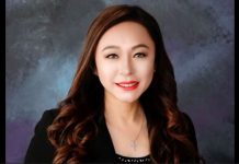 Professional headshot of a smiling Asian woman with long wavy brown hair, wearing a black blazer and necklace, against a dark textured backdrop.