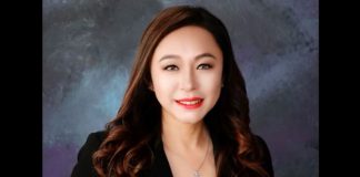 Professional headshot of a smiling Asian woman with long wavy brown hair, wearing a black blazer and necklace, against a dark textured backdrop.