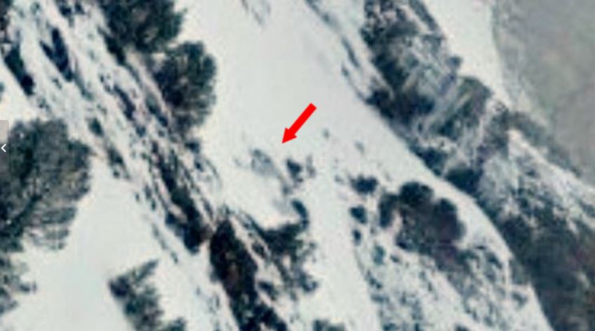 Aerial view of a snow-covered mountain slope with a red arrow pointing to a small climber or object on the slope near rocks.