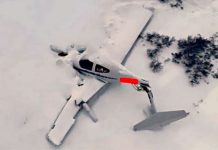 Propeller airplane wreck in deep snow with the fuselage partially buried and red marking on the side, in a snowy landscape.