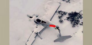 Propeller airplane wreck in deep snow with the fuselage partially buried and red marking on the side, in a snowy landscape.
