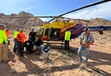 Rescue crew gathers around a yellow helicopter on a rocky desert site as they assist a person on the ground.