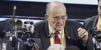 Older man in a suit and red tie speaks to reporters amid cameras and microphones at a press event.