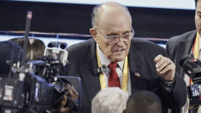 Older man in a suit and red tie speaks to reporters amid cameras and microphones at a press event.