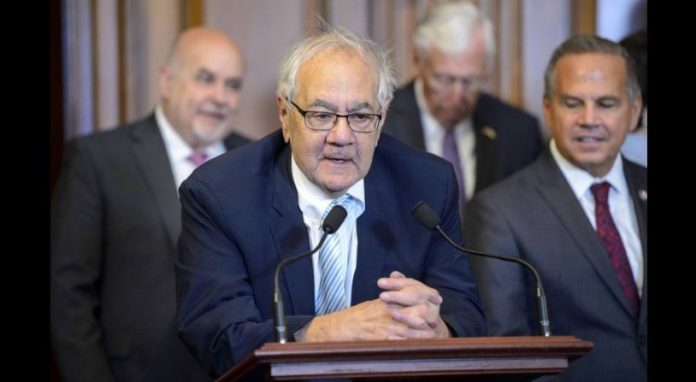 Man in a navy suit and glasses speaks at a wooden podium with two microphones, flanked by suited men in a formal setting.