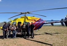 Group of eight people posing in front of a bright yellow rescue helicopter with red and purple stripes on a dry, sunny field.