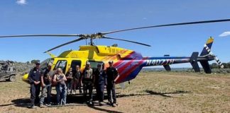 Group of eight people posing in front of a bright yellow rescue helicopter with red and purple stripes on a dry, sunny field.