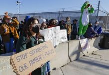 Protesters behind a chain-link fence hold signs, including 'Stop separating our families' during a daytime demonstration.