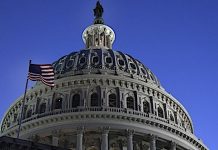 US Capitol dome with an American flag waving in the foreground against a clear blue sky