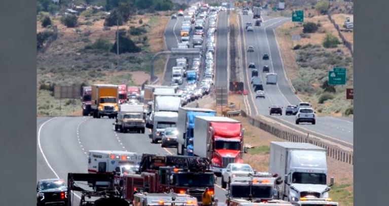 Long line of vehicles, including many trucks, stuck in heavy traffic on a sunny multi-lane highway.