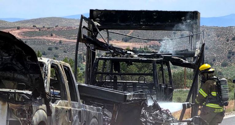 Firefighter in a yellow helmet spraying water on charred vehicles after a bus fire in a rural landscape.