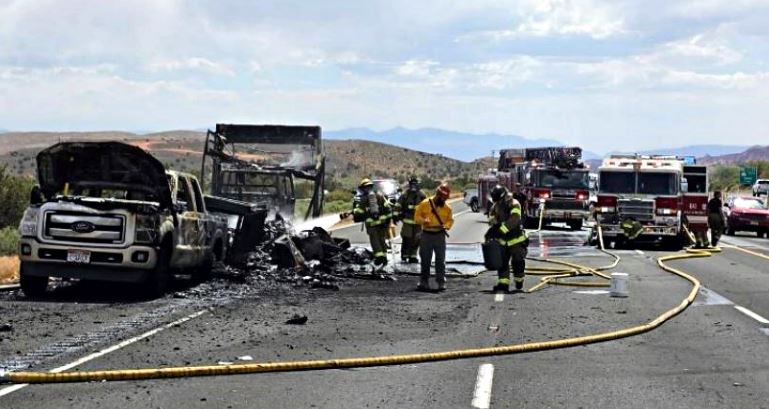 Firefighters in turnout gear spraying water on a burned truck and debris on a highway, with fire engines and hoses in the background.