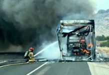 Firefighter spraying water on a burned bus on a highway with heavy smoke and mountains in the background.