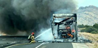Firefighter spraying water on a burned bus on a highway with heavy smoke and mountains in the background.