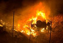 Car is engulfed in flames at night behind a wire fence, a vehicle burning intensely on a hillside campground or field area.