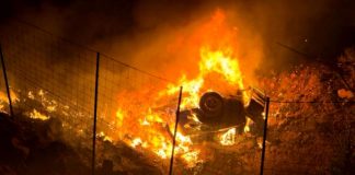 Car is engulfed in flames at night behind a wire fence, a vehicle burning intensely on a hillside campground or field area.
