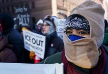 Protester with a beige balaclava and hood, at a rally, holding a sign calling to 'ICE OUT' near the foreground.
