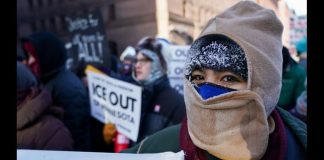 Protester with a beige balaclava and hood, at a rally, holding a sign calling to 'ICE OUT' near the foreground.