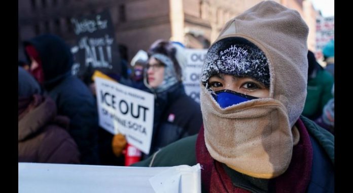 Protester with a beige balaclava and hood, at a rally, holding a sign calling to 'ICE OUT' near the foreground.
