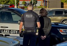 Police officer in tactical vest stands beside a handcuffed person next to a Salt Lake City police SUV in a parking lot.