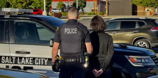 Police officer in tactical vest stands beside a handcuffed person next to a Salt Lake City police SUV in a parking lot.