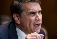 Close-up of a man in a dark suit speaking, gesturing with his hand in a formal setting.