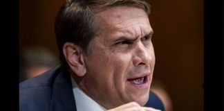 Close-up of a man in a dark suit speaking, gesturing with his hand in a formal setting.