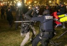 Police officers in tactical gear restrain a protester at night while photographers and bystanders film the scene in a park-like area.