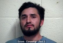 Mugshot of a man with dark hair and beard standing in front of a light cinder block wall; caption reads Iron County Jail.