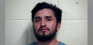 Mugshot of a man with dark hair and beard standing in front of a light cinder block wall; caption reads Iron County Jail.