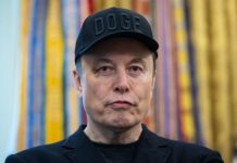 Person wearing a black cap with 'DOGE' lettering, dark blazer, neutral expression, colorful striped background.