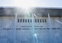 Facade of a light-colored building with tall columns and an inscription reading 'The Donald Trump and The John F. Kennedy Memorial Center for the Performing Arts,' with sun glare at the top.