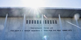 Facade of a light-colored building with tall columns and an inscription reading 'The Donald Trump and The John F. Kennedy Memorial Center for the Performing Arts,' with sun glare at the top.