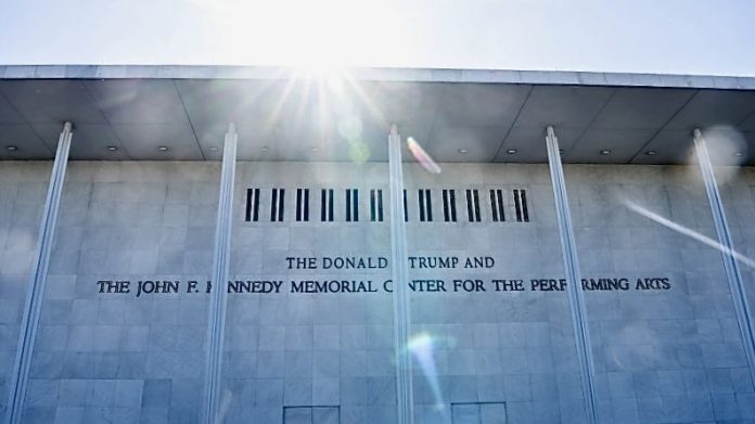 Facade of a light-colored building with tall columns and an inscription reading 'The Donald Trump and The John F. Kennedy Memorial Center for the Performing Arts,' with sun glare at the top.
