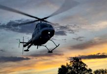 A helicopter in flight over trees at sunset, with rotor blur and landing skids visible against a colorful sky.