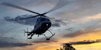 A helicopter in flight over trees at sunset, with rotor blur and landing skids visible against a colorful sky.