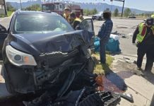 Severely damaged dark SUV with a crushed front end at a roadway crash scene, with emergency responders and a fire truck in the background.