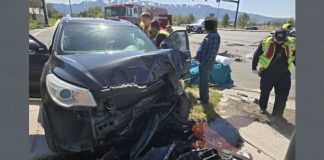 Severely damaged dark SUV with a crushed front end at a roadway crash scene, with emergency responders and a fire truck in the background.