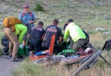 Emergency responders kneel around a fallen cyclist on a dirt path, tending to an injured rider with a toppled bike nearby in a grassy hillside setting.