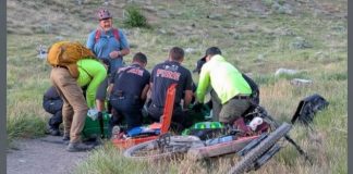 Emergency responders kneel around a fallen cyclist on a dirt path, tending to an injured rider with a toppled bike nearby in a grassy hillside setting.