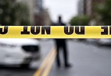 Yellow caution tape stretches across a street, with a blurred figure and a white car in the background (crime scene barrier).