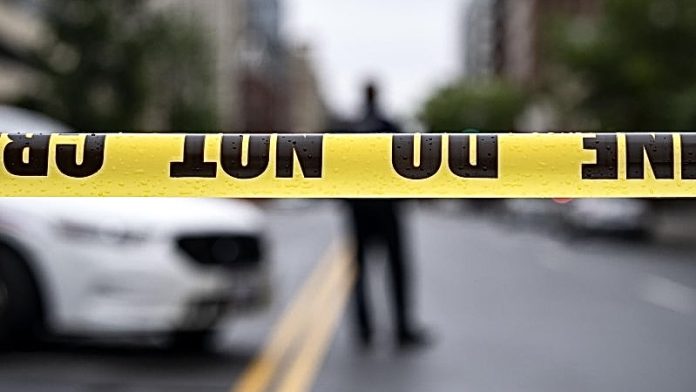 Yellow caution tape stretches across a street, with a blurred figure and a white car in the background (crime scene barrier).