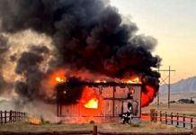 House on fire: a small wooden structure engulfed in flames with thick black smoke, firefighters near the front as the roof burns away.