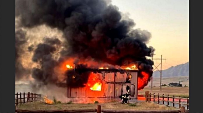 North Tooele Fire 1 House on fire: a small wooden structure engulfed in flames with thick black smoke, firefighters near the front as the roof burns away.